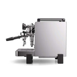 A modern espresso machine with a brushed silver metal body, black accents, and a black steam wand handle, shown from a three-quarters angle on a white background.